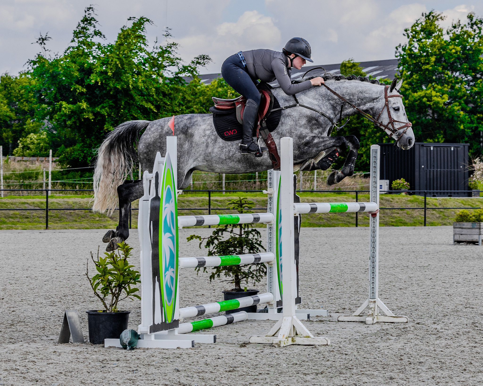 A person wearing a grey long sleeve base layer leaning on a metal fence wearing white equestrian breeches and a grey cap