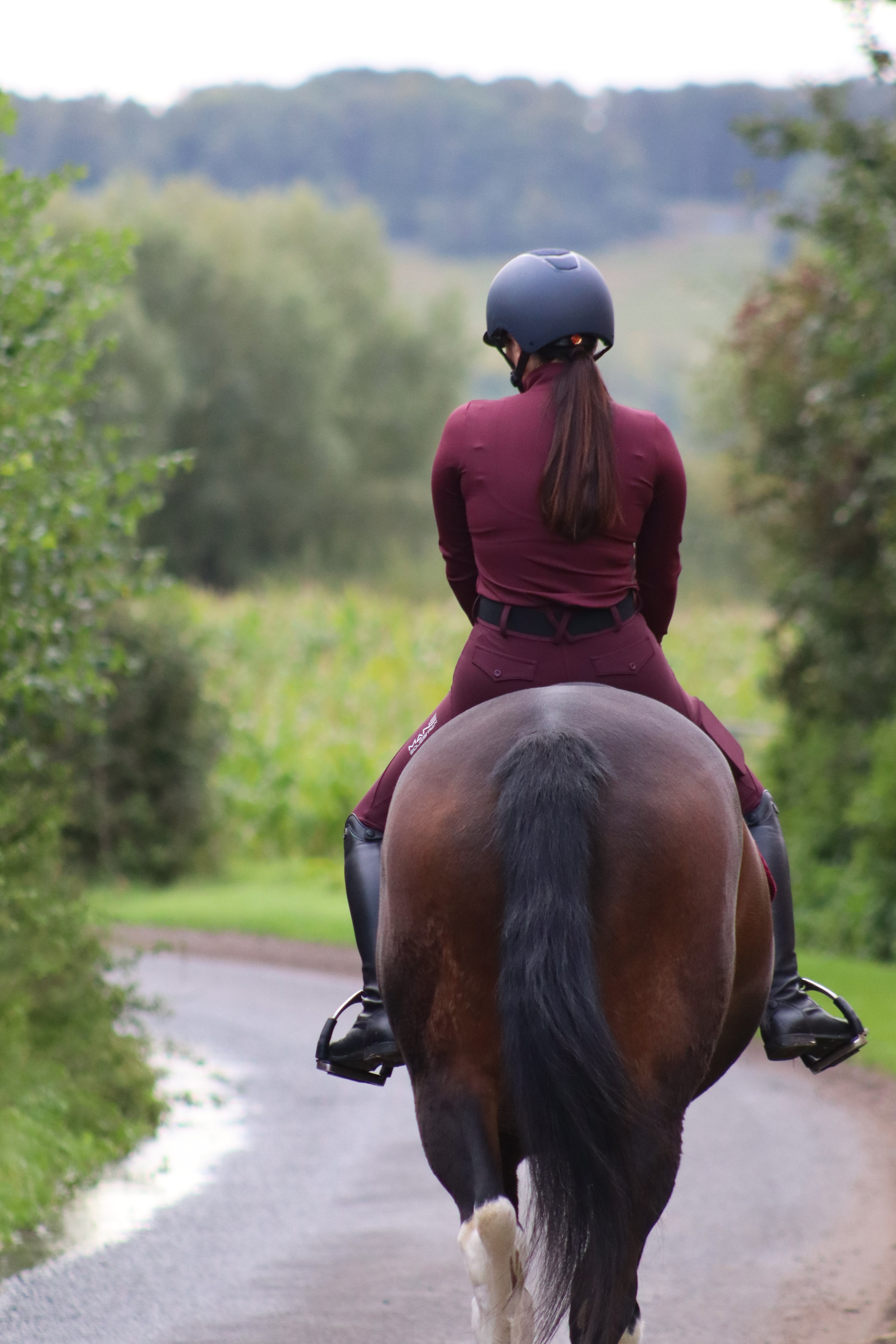 A person stood next to an apple tree wearing burgundy long sleeved performance base layer and matching riding breeches with a logo on the baselayer chest
