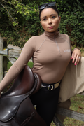 A person wearing a brown turtleneck top and a white belt with a silver snaffle design holding on a brown saddle.