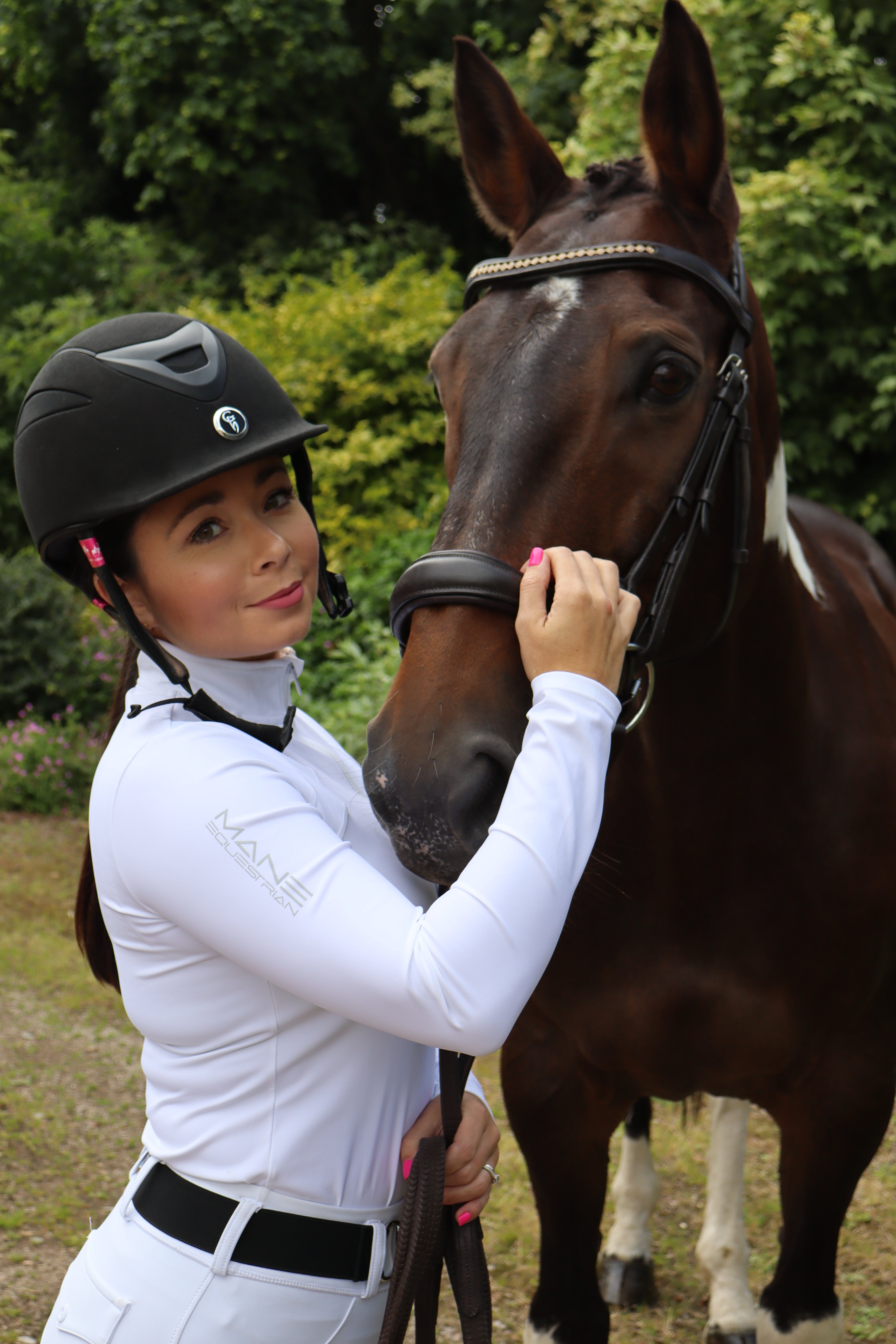 A person wearing a white performance base layer top with reflective details, standing next to a black and white horse. The person is holding the horse's bridle.