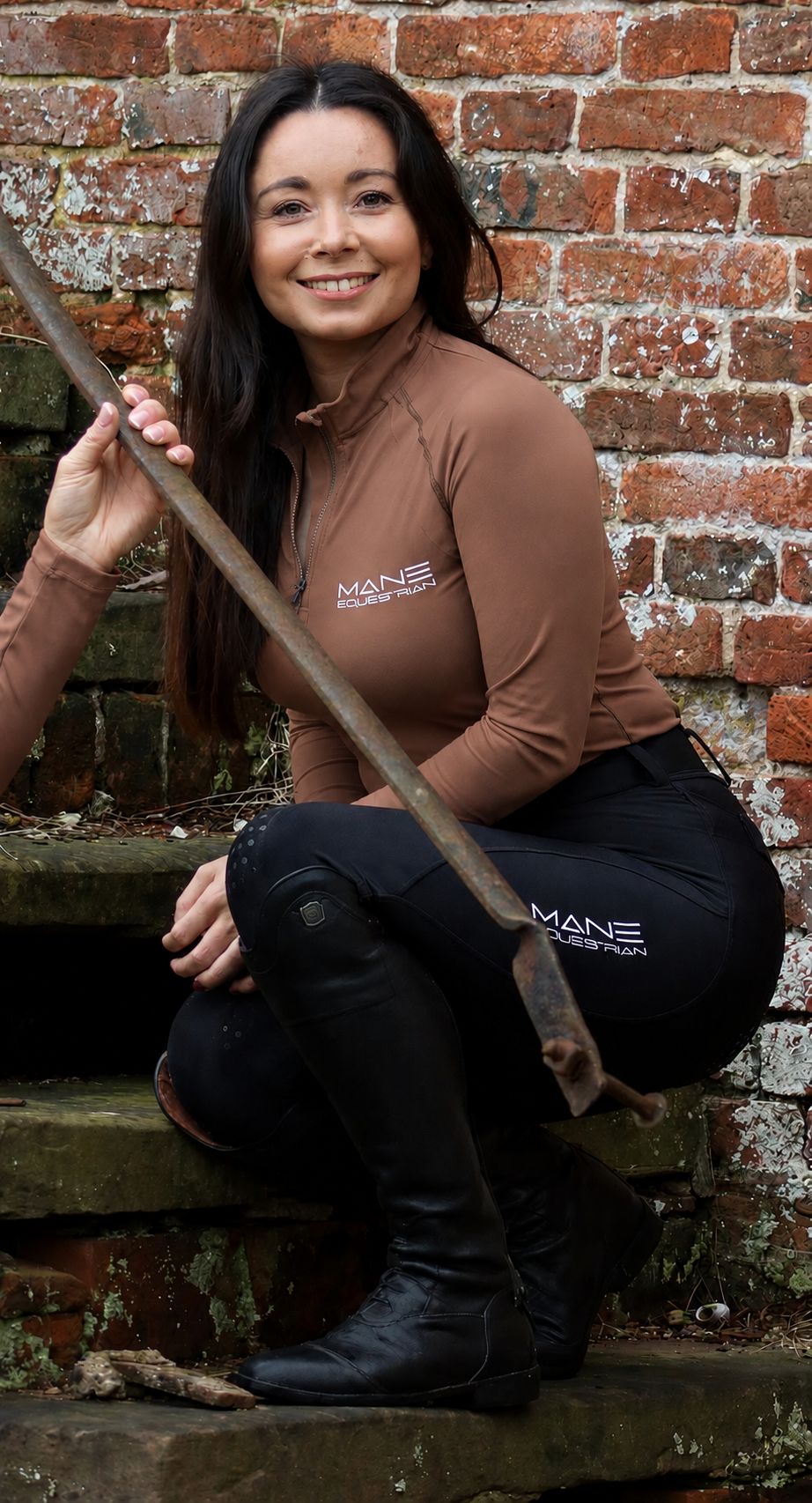 A person wearing a chestnut baselayer with black performance breeches sat on steps against a brick wall
