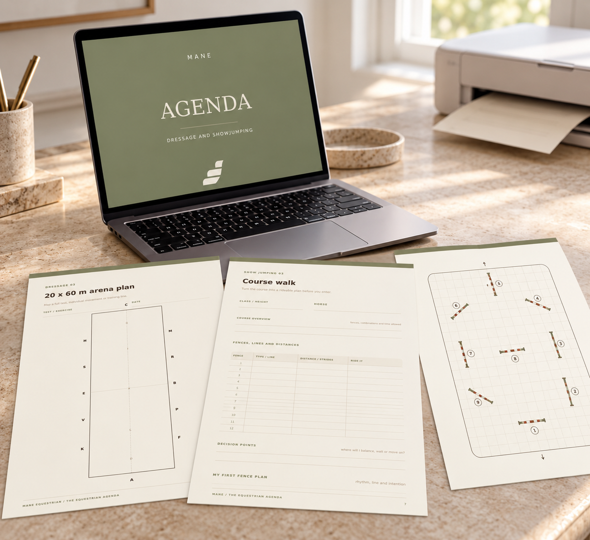 A laptop on a sunlit desk displays an agenda screen, with printed agenda sheets spread out in front. The scene includes a printer, cup with pens, and a window in the background, creating a styled workspace setup.